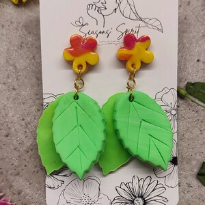 May include: A pair of earrings featuring a green leaf design with a yellow, orange, and pink flower charm. The earrings are displayed on a white card with the text "Seasons' Spirit Costa del Maresme".
