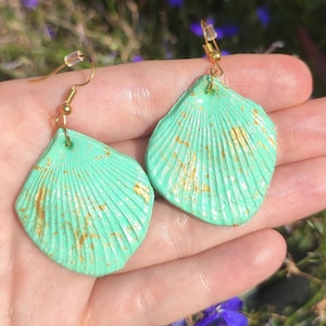 May include: A pair of seafoam green seashell-shaped earrings with gold accents. The earrings have a textured surface with vertical lines and gold flecks. They are suspended from gold-colored ear wires, showcasing a coastal theme.