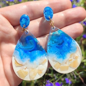 May include: A pair of teardrop-shaped resin earrings with a blue and white marbled design. The earrings have a gold-colored metal hook and are filled with small white pieces.