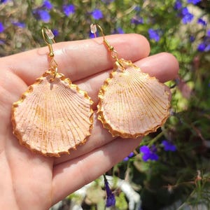 Op de afbeelding: Een paar gouden oorbellen in de vorm van schelpen met een roze en wit ontwerp. De oorbellen hebben een delicate, vrouwelijke uitstraling.