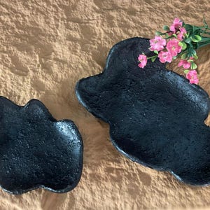 May include: Two black, irregularly shaped, ceramic dishes. The larger dish is on the right and the smaller dish is on the left. The dishes are on a light brown textured surface. Pink flowers are in the background.