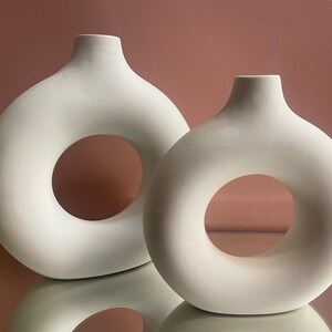 May include: Two white ceramic vases with a donut-shaped opening. The vases are different sizes and are sitting on a reflective surface.