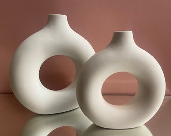 Może przedstawiać: Dwa białe ceramiczne wazony z otworem w kształcie pączka. Wazony są różnych rozmiarów i stoją na odbijającej powierzchni.