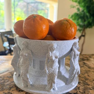 May include: A decorative fruit bowl filled with bright orange tangerines. The bowl is light gray with a speckled texture and features sculpted figures of women around its base. The bowl is a unique piece for displaying fruit.