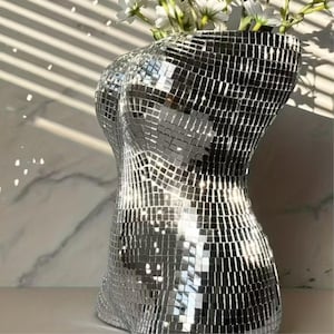 May include: A decorative vase in the form of a torso, covered in small, reflective silver mosaic tiles. The vase holds white flowers. The mosaic design creates a shimmering effect, reflecting light.