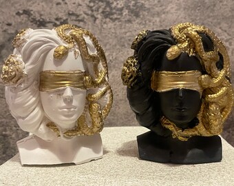 May include: Two Medusa bust sculptures, one white and one black, each with gold accents. The busts feature a blindfold and snakes in their hair. The sculptures are decorative and artistic.