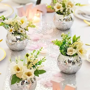 May include: Four small, round, silver mosaic vases filled with white and yellow flowers and greenery. The vases are arranged on a silver glitter table runner, with scattered pink petals and candles. The setting is a festive, celebratory theme.