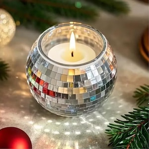 May include: A spherical glass candle holder with a mosaic mirrored exterior. A lit white candle sits inside. The candle flame is yellow. The background includes a red bauble and green fir needles.