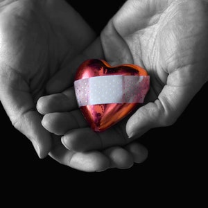 Può includere: Un oggetto rosso a forma di cuore con una benda bianca avvolta intorno, tenuto nelle mani di una persona.