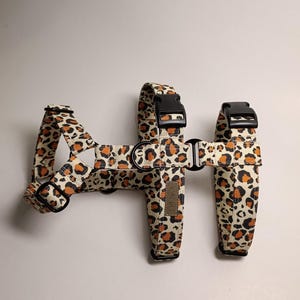 May include: A pet harness featuring a beige fabric with a black and orange leopard print pattern. The harness includes black plastic buckles, adjustable sliders, and metal D-rings for leash attachment. A small rectangular brand label is visible.
