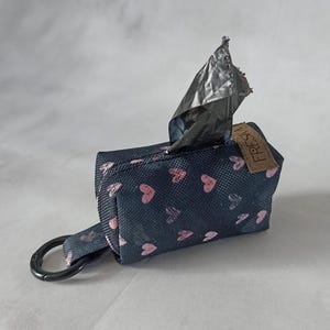 May include: A black and pink dog waste bag dispenser with a heart pattern. The dispenser has a black metal ring for attaching to a leash. The dispenser is labeled "FRESH".