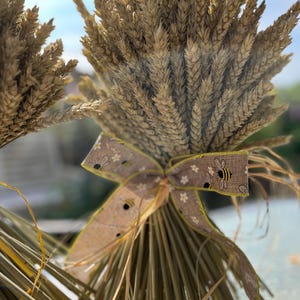 May include: A bunch of dried wheat stalks tied with a decorative ribbon. The ribbon is tan with a pattern of small white flowers and bees. The wheat is a light brown colour.