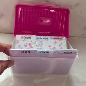 以下が含まれることがあります： 鮮やかなピンク色の蓋が付いた透明なプラスチック製の収納ボックス。中には整理用の仕切りが入っています。仕切りには花柄が施されており、SUN、MON、TUE、WED、NEXT WEEK、COMPLETEというラベルが付けられています。長方形のヒンジ付き蓋のデザインです。