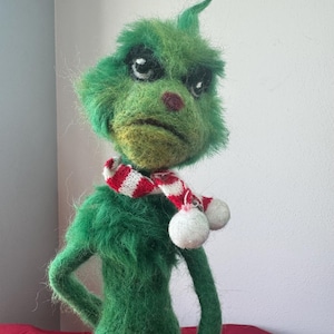 Op de afbeelding: Een naaldgevilte groene wezenfiguur met een textuurrijk, pluizig uiterlijk, grote donkere ogen, een kleine rode neus en een neerwaarts gerichte mond. Het figuur draagt een rood-wit gestreepte gebreide sjaal met twee witte pompons.