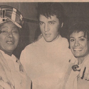 May include: A black and white newspaper clipping with a photo of Elvis Presley, a woman, and another woman. The headline reads "Psychiatrist Elvis!" and the article discusses Elvis's latest film, "A Change of Habit". The article also includes a quote from the woman in the photo, who says "I finally met the man - the King - Elvis Presley!"