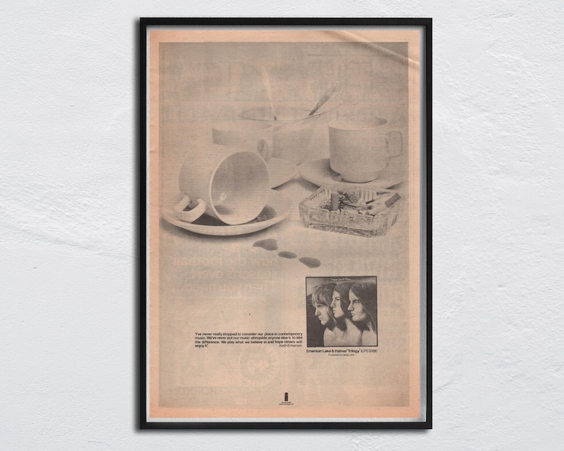May include: Framed vintage print featuring a still life with teacups, saucers, and an ashtray. The print has a sepia tone and includes text and a black and white photo of three people. The frame is black.