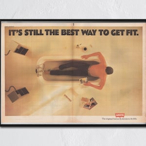 以下が含まれることがあります： リーバイスジーンズの額入りヴィンテージ広告。画像には、バスタブに入っている人物と「IT'S STILL THE BEST WAY TO GET FIT」というテキストが表示されています。リーバイスのロゴは下部にあり、追加のテキストも表示されています。