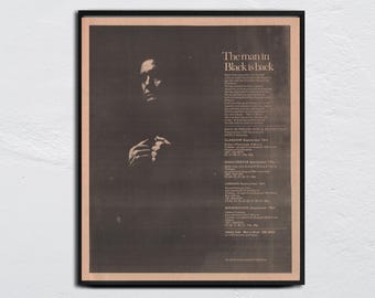 May include: A framed vintage poster with a sepia-toned image of a man in a dark suit. The text "The man in Black is back" is at the top, with tour dates listed below. The poster is in a black frame.