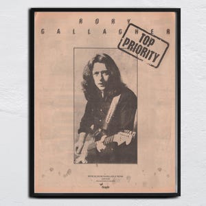 Può includere: Poster vintage incorniciato con Rory Gallagher che suona la chitarra. L'immagine in bianco e nero è ambientata su uno sfondo beige con testo in alto e in basso. Un timbro recita "TOP PRIORITY". Il poster promuove un nuovo album.