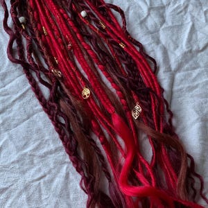Könnte beinhalten: Ein Set langer, roter und burgunderfarbener synthetischer Dreadlocks mit goldenen Blattakzenten und Perlen. Die Dreadlocks sind oben mit einem Gummiband zusammengebunden.