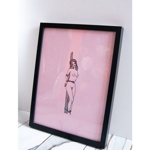 May include: Framed art print featuring a black and white illustration of a person tied to a wooden post against a pink background. The artwork is encased in a black frame, creating a striking contrast.