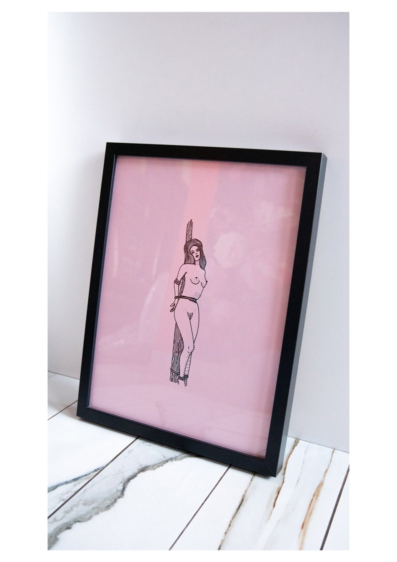 May include: Framed art print featuring a black and white illustration of a person tied to a wooden post against a pink background. The artwork is encased in a black frame, creating a striking contrast.
