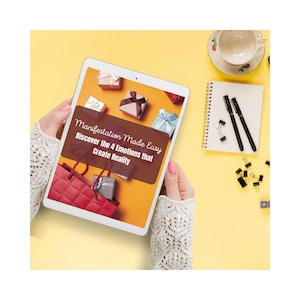 May include: A tablet displaying the text "Manifestation Made Easy" with gift boxes and a red shopping bag on a yellow background. A notepad, pens, paper clips, and a teacup are also visible.