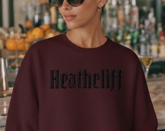 May include: A burgundy sweatshirt with the word "Heathcliff" embroidered in black gothic lettering. The sweatshirt is a solid color and has a crew neck. The person in the image is wearing sunglasses and gold hoop earrings.