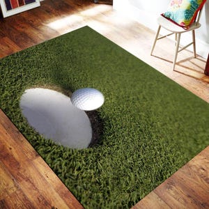 Könnte beinhalten: Ein rechteckiger Teppich mit einem hochauflösenden Fotodruck eines Golfplatz-Greens. Das Design zeigt eine texturierte grüne Grasfläche mit einem weißen Sandbunker und einem einzelnen weißen Golfball, der über dem Sand schwebt.