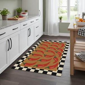 May include: A rectangular kitchen rug featuring a repeating pattern of red chili peppers on a tan background. The rug has a black and white checkered border. The rug is placed on a dark wood floor in front of white kitchen cabinets.