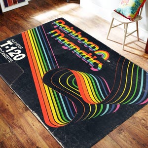 May include: A rectangular area rug featuring a black background with a graphic design of a vintage VHS cassette tape label. The design includes rainbow-colored stripes and the text Rainbow Magnetics, T-120, 2-4-6 Hour, and Video Cassette System.