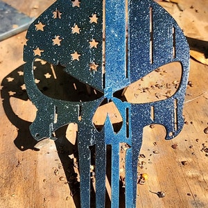 Puede incluir: Una silueta de cráneo de metal con un acabado de pintura azul y negro. El cráneo está dividido en dos, con un lado que presenta un diseño de línea azul fina azul y blanco y el otro lado que presenta un diseño de bandera estadounidense azul y negro.