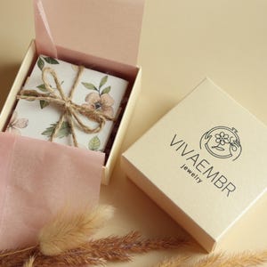 May include: A white box with a floral design and a brown twine bow. The box is open and the inside is lined with pink tissue paper. The box is sitting on a beige surface. The box is labelled "Vivaembr Jewelry".