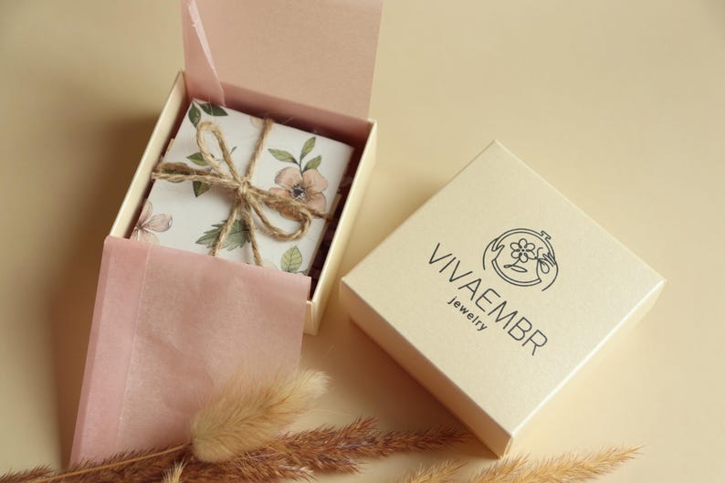 May include: A white box with a floral design and a brown twine bow. The box is open and the inside is lined with pink tissue paper. The box is sitting on a beige surface. The box is labelled "Vivaembr Jewelry".