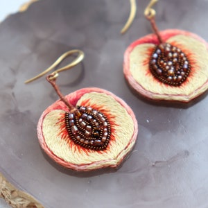 May include: A pair of peach-shaped earrings with embroidered details and brown seed beads. The earrings are made of fabric and have gold-colored hooks.