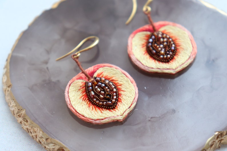 May include: A pair of peach-shaped earrings with embroidered details and brown seed beads. The earrings are made of fabric and have gold-colored hooks.