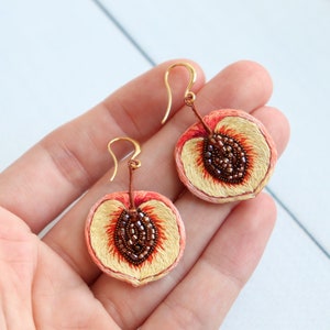 May include: A pair of peach-shaped earrings with gold hooks. The peaches are embroidered with peach fuzz and have a brown seed detail.