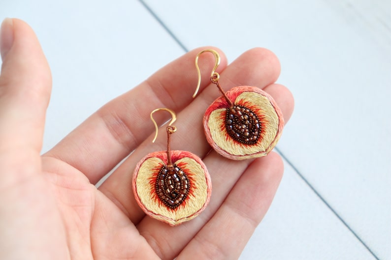 May include: A pair of peach-shaped earrings with gold hooks. The peaches are embroidered with peach fuzz and have a brown seed detail.