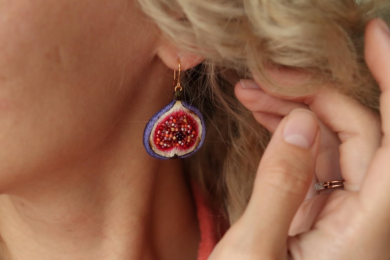 May include: A pair of earrings shaped like a halved fig with a purple outer layer and a red and white interior. The earrings are made of fabric and beads.