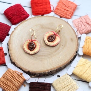 May include: A pair of peach-shaped earrings with gold hooks. The earrings are embroidered with peach slices and have brown bead accents. The earrings are displayed on a wooden surface with spools of embroidery floss in various shades of orange and red.
