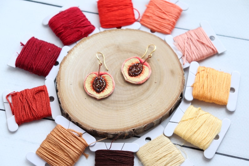 May include: A pair of peach-shaped earrings with gold hooks. The earrings are embroidered with peach slices and have brown bead accents. The earrings are displayed on a wooden surface with spools of embroidery floss in various shades of orange and red.