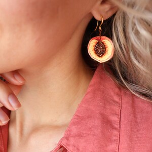 May include: Gold dangle earrings with a peach slice design. The peach is painted with realistic details, including a yellow flesh, red skin, and brown seeds.