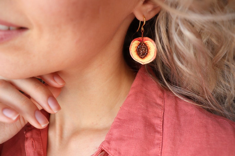 May include: Gold dangle earrings with a peach slice design. The peach is painted with realistic details, including a yellow flesh, red skin, and brown seeds.