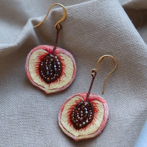 May include: A pair of peach-shaped earrings with gold hooks. The earrings are embroidered with peach slices and have a brown seed detail.