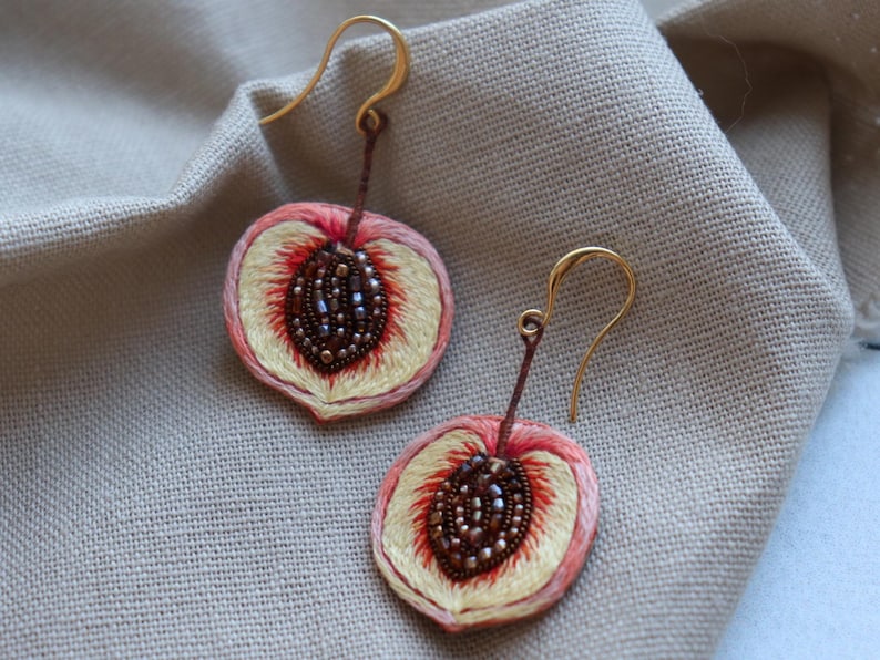 May include: A pair of peach-shaped earrings with gold hooks. The earrings are embroidered with peach slices and have a brown seed detail.