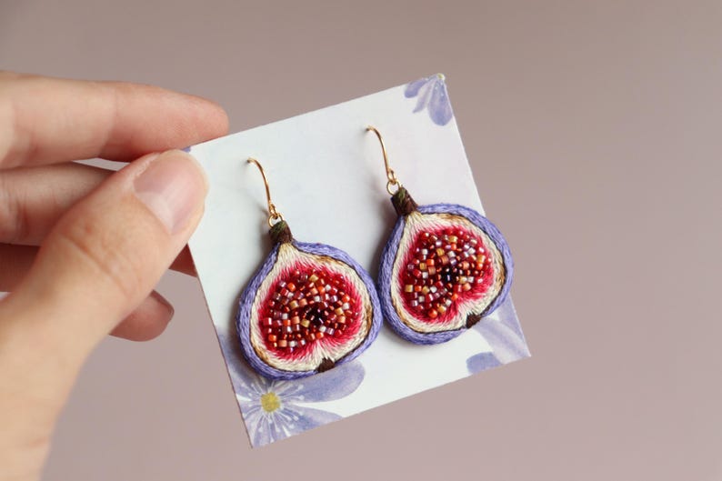 May include: A pair of dangle earrings featuring a detailed embroidered fig design. The figs are a vibrant purple with a red and pink interior. The earrings are made with gold-toned metal.