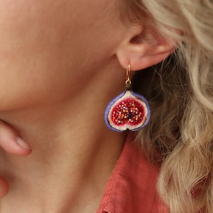 May include: A pair of dangle earrings featuring a purple fig with a red interior and white seeds. The earrings have gold hooks.