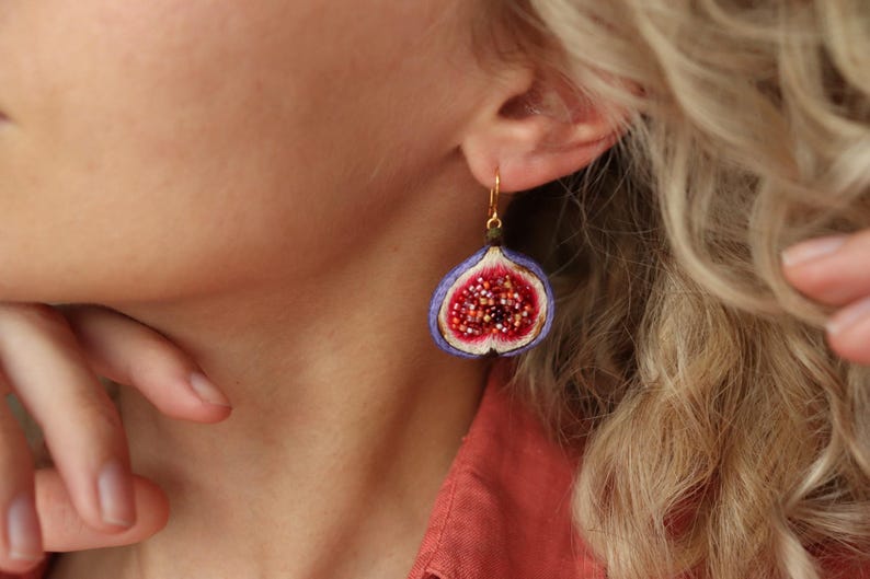 May include: A pair of dangle earrings featuring a purple fig with a red interior and white seeds. The earrings have gold hooks.