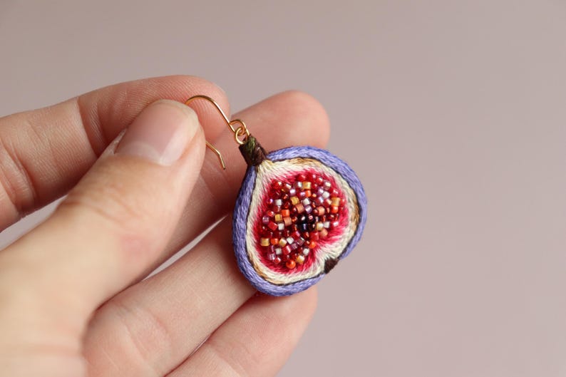 May include: Purple and white fig earrings with a gold hook. The fig is embroidered with red and orange beads.