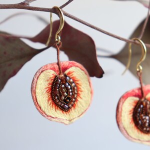 May include: A pair of peach-shaped earrings with embroidered details. The peaches are a light peach color with a darker peach center and brown seed details. The earrings are hanging from gold hooks.